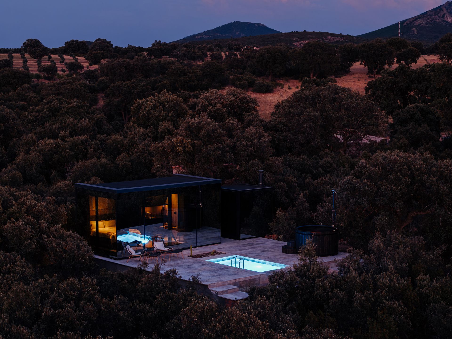 Casa Espejo de Cork Valley iluminada al anochecer, con piscina propia y jacuzzi, rodeada de encinas: alojamiento discreto para parejas cerca de Madrid